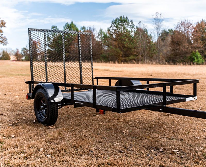 Black metal utility trailer with mesh ramp gate parked on a grassy field with trees in the background.
