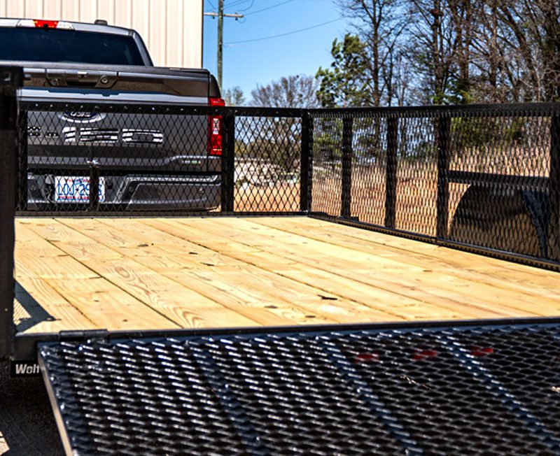View from the back of a utility trailer with a wooden deck and metal mesh sides, hitched to a Ford pickup truck with a visible license plate.