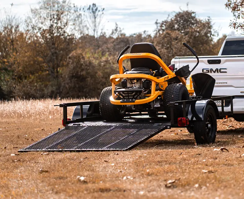 Yellow riding lawn mower being loaded onto a black utility trailer with a lowered mesh ramp, attached to a white GMC pickup truck in a grassy area.