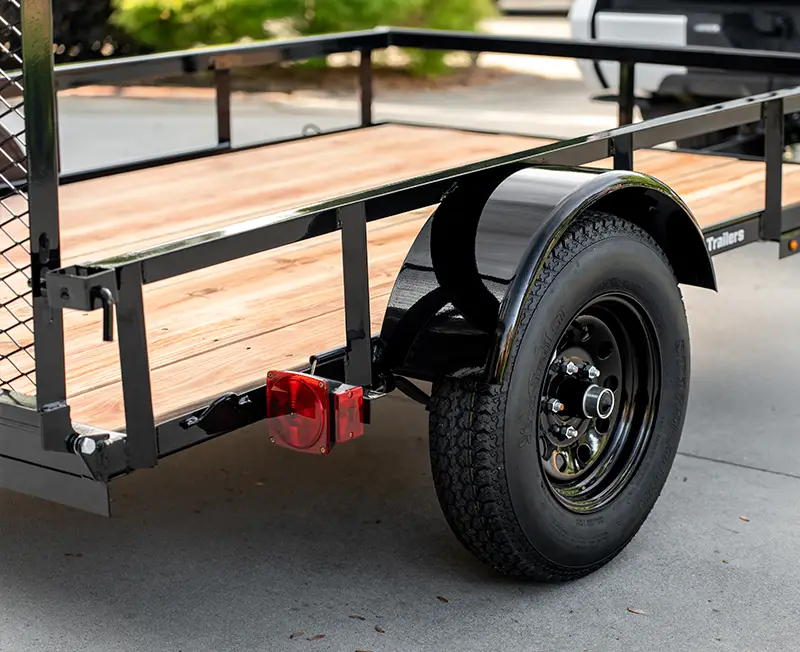 Close-up of a utility trailer with a wood plank deck, black steel frame, fender, wheel, and rear taillight parked on a driveway.