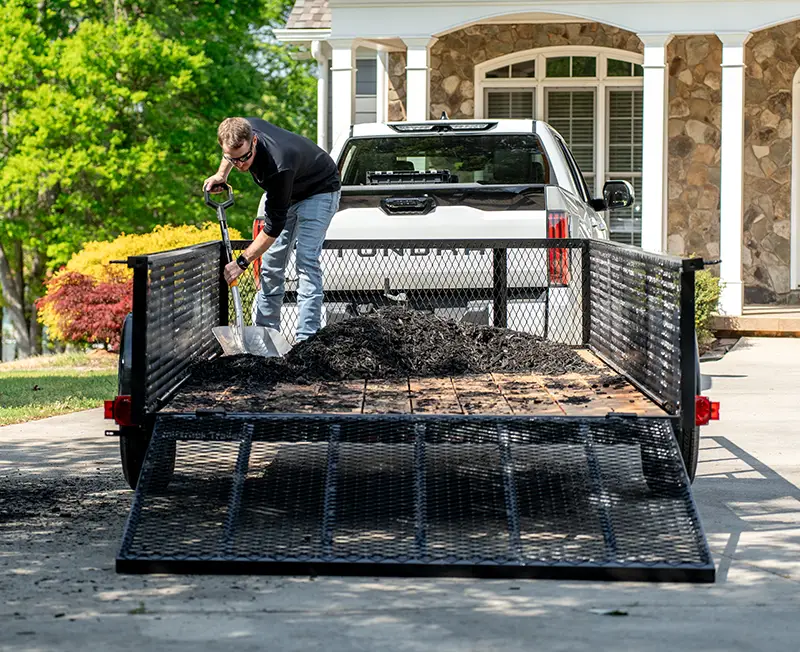 Person shoveling mulch from a utility trailer with mesh sides and a fold-down ramp, attached to a pickup truck in a residential driveway.