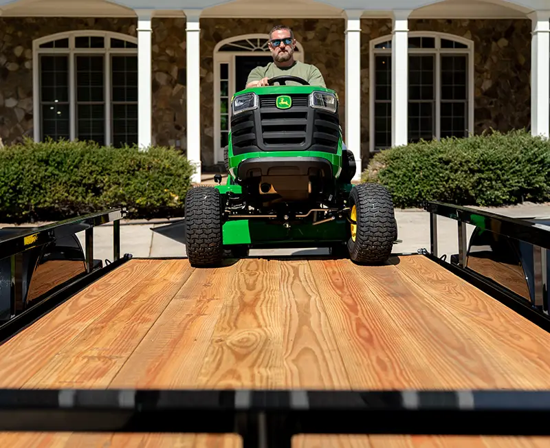 Riding lawn mower being driven onto a utility trailer with a wood deck in front of a house.