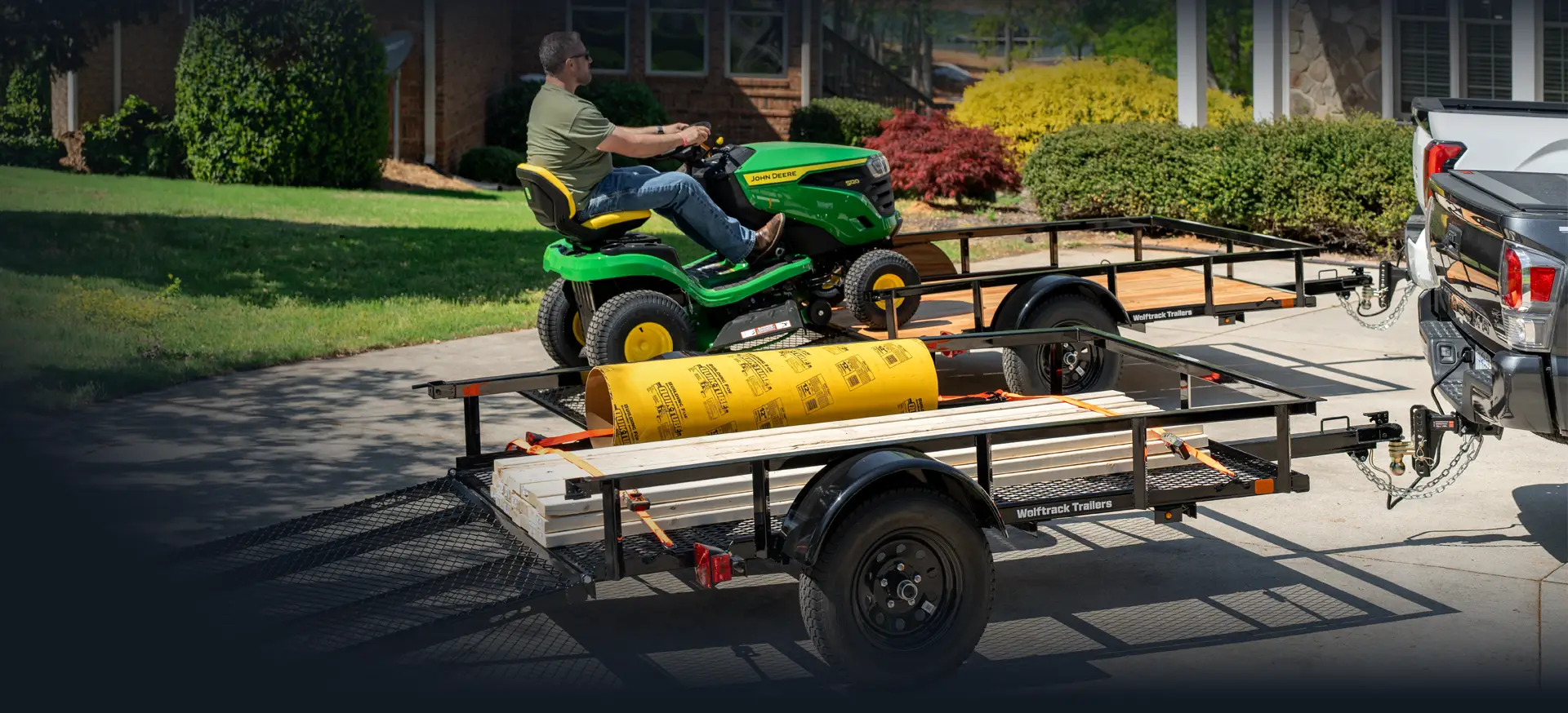 Two utility trailers hitched to a pickup truck; one carries a riding lawn mower and the other is loaded with lumber and a roll of material secured with straps.
